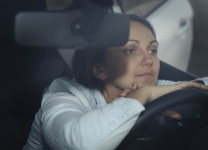 Participate - View through the windscreen of a woman sitting waiting in her car with hands and chin resting on the steering wheel